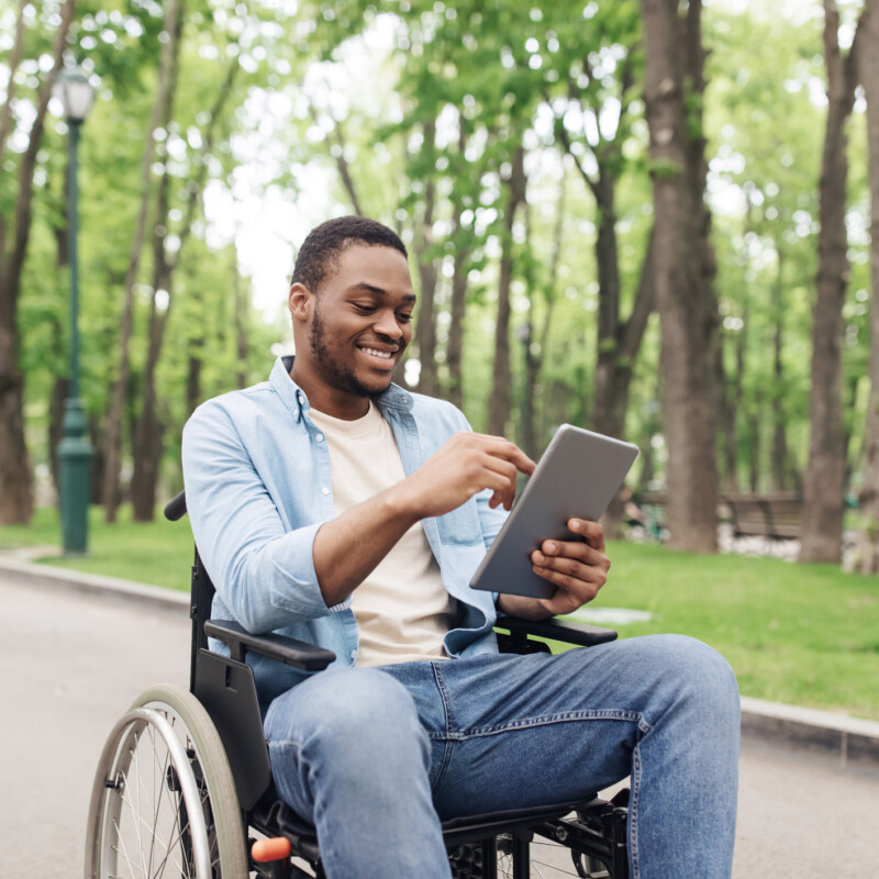 Happy young black man in wheelchair using tablet pc, browsing web or watching movie at spring park. Millennial African American guy checking social media on mobile device outdoors